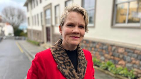 A woman with short blonde hair looking at the camera and smiling. She is wearing a red coat with a leopard-print collar.