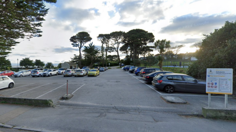 Gyllyngvase Coach Park in Falmouth is around two thirds full of cars. There are old pine trees to the left and back of it and to the right is a park. A sign in the foreground has the parking tariff but it is too far away to read what it says except for Cornwall Council.  