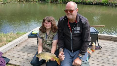 Shona and Mr Barker sitting near water while holding a fish. Shona is wearing red-rimmed glasses and has brown hair.