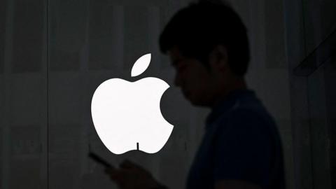 The Apple logo in white against a black background. There is a silhouette of someone holding a mobile phone