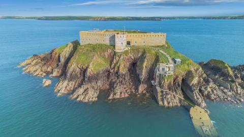 An aerial view of Thorne Island, with the yellowed fortress walls encompassing it and cliffs covered in greenery at its edges. The island is surrounded by the sea, with the mainland in distance. Two flights of steps lead down to the water's edge. 