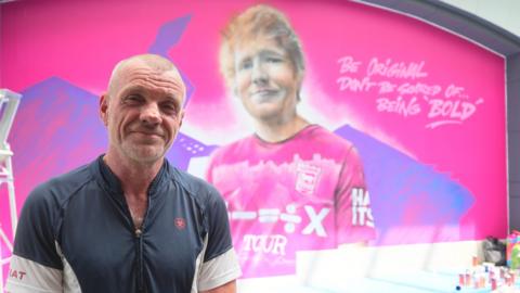 Keith Hopewell a man who is looking directly at the camera and smiling. He is standing inside a shopping centre in front of a bright pink wall which has a mural of Ed Sheeran on. 