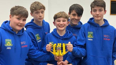 A group of five boys all in blue hoodies holding a trophy made of Lego. They have mainly brown or dark hair and are all smiling.