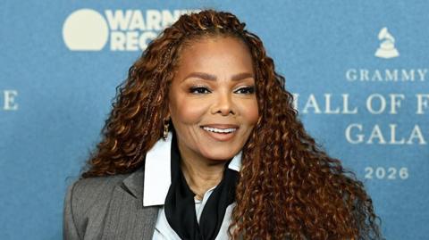 Smiling Janet Jackson stood at a press event against a blue backdrop
