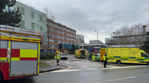 A fire engine and several ambulances are parked up on roads around Southampton General Hospital. A police officer can be seen standing at the end of the road leading to the hospital. Several other emergency service personnel can be seen walking around the scene in hi-viz clothing. 