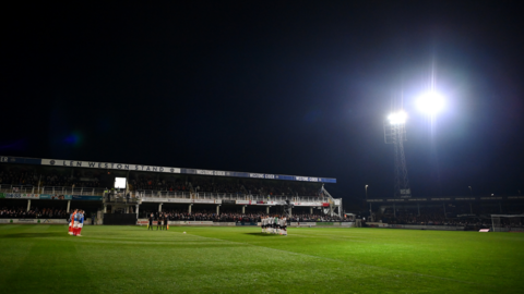 A grassy pitch with football stands and a floodlight in the distance, seen at night