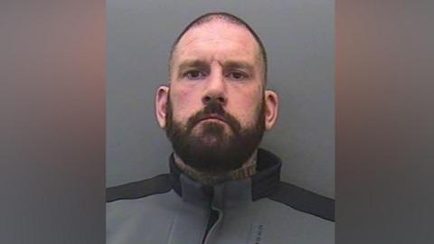 Gary Parkinson looking at the camera. He has short black hair and a full beard. He is wearing a black and grey tracksuit jumper.