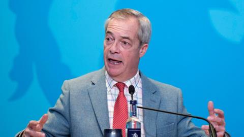 Nigel Farage sat at a desk in front of a blue backdrop, gesticulating with his hands. He is wearing a grey suit, red tie with white spots and a chequered shirt.