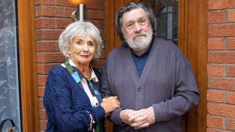 Sue Johnston and Ricky Tomlinson are pictured in character, standing in front of a semi-detached red-bricked house on Brookside Close. A wooden door with a glass panel is behind them and a window is to the left. Johnston is wearing a colourful patterned dress and a navy cardigan and Tomlinson is wearing a navy polo shirt and dark grey cardigan.