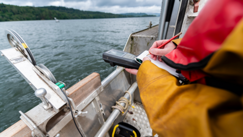 A scientist - who wears yellow overalls and a red life jacket - records some figures from a machine and writes them down with a pencil on paper. They are standing on the side of a boat which is sailing on Windermere.