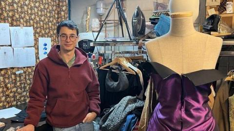 Cosmin Diaconu is standing on the left of his studio. He has short brown hair and is wearing a red jumper and jeans. On the right is a mannequin which is wearing a purple dress.