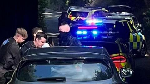 Two police officers in black uniforms standing to the left of a black car. Behind the black car are two police cars, with blue-and-yellow markings and blue lights shining. 