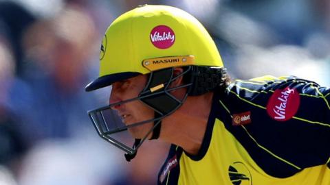 Hampshire Hawks batter Hilton Cartwright running across a wicket during a T20 Blast match against Kent Spitfires while wearing a predominantly yellow kit and helmet with dark blue, approaching an opposition player wearing a blue and red kit
