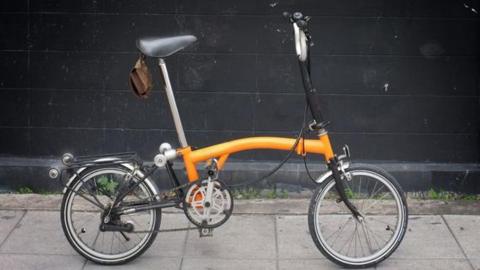 An orange Brompton folding bicycle is on a pavement next to a wall.
