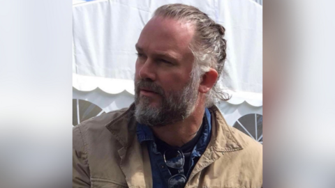 A man with a grey beard and hair tied back looking to the side. He is wearing a beige jacket and a blue shirt with sunglasses hanging on.