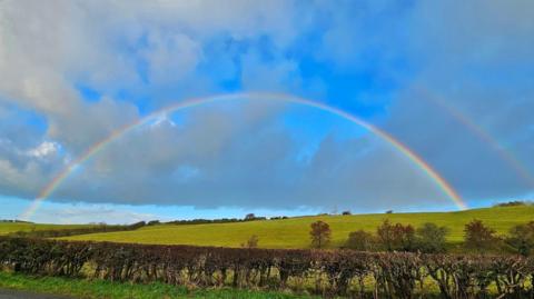 A rainbow over some green fields, a fainter rainbow can be seen above it. The sky is blue with white and grey clouds. There is a hedge in the foreground and trees in the field.