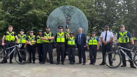 Several male and female police officers in unform stand with Simon outside by a circular statue. Two officers are standing with bikes.
