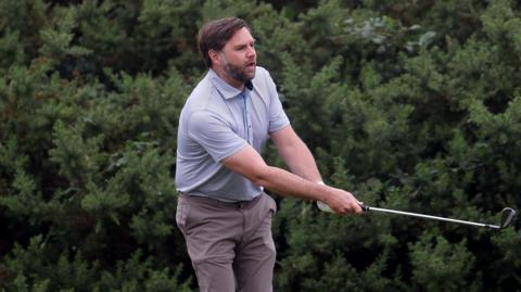 A man with brown hair and beard, wearing a light blue top an brown trousers swings a golf club with bushes behind him.