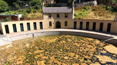 A drone image of Cleveland Pools, showing dark water with patches of what looks like yellow/brown algae. The Cotswold stone-style buildings are visible in the background