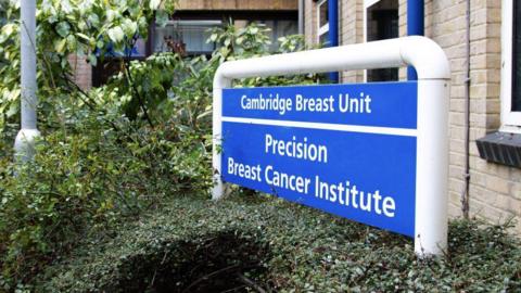 A sign outside of a building is on the left. It is blue and has white writing on it, which reads "Cambridge Breast Unit Precision Breast Cancer Institute". There are hedges and trees surrounding the sign. 