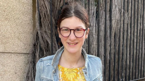 Anna Beaumont wearing a blue denim jacket and a yellow top. She is wearing glasses with her hair tied back. She is smiling at the camera. 