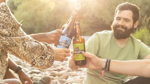 Four people sit on a pebble beach by a river, clinking their bottles of Ilkley Brewery beer 