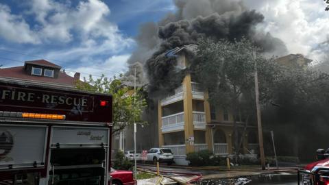 Dark grey smoke billows from a multi-story building. Multiple fire vehicles are seen nearby with hoses draped on the street where multiple cars are parked. 