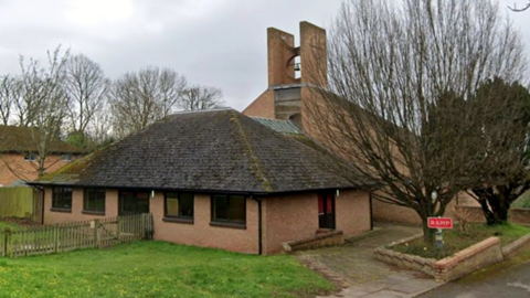 A church building with grass and a fence in the foreground. Trees are on the left and right of the image.