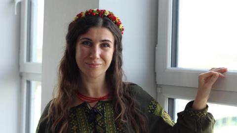 A woman with long brown hair stands by a window in the daytime, smiling into the camera. She wears a dark green traditional Ukrainian blouse with embroidery on the front and sleeves, and a headband decorated with small red and yellow flowers.