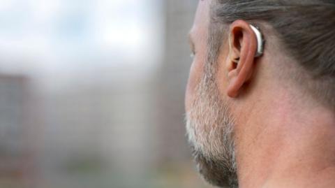 Stock image of man with a grey beard wearing a hearing aid. The shot, taken from behind, shows the anonymous man's left ear. The hearing aid is a silver-coloured, sleek modern device tucked behind his earlobe. 