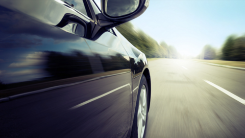 An image of the front-right of a dark-coloured car as it drives along a road.