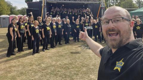 A man in a black polo taking a selfie with lots of choir members on a stage and off stage in the background. They are at a festival.