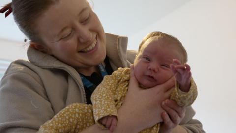 Midwife Hannah holding a newborn baby, smiling. The baby, dressed in a yellow outfit with white floral patterns, is gazing at the camera with one hand raised. 