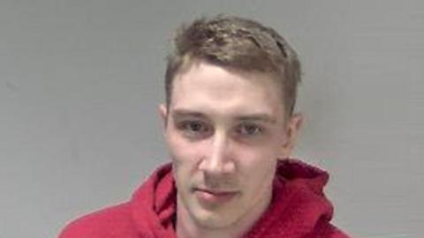 A mugshot of a man in a red hoodie looking at the camera in front of a grey background