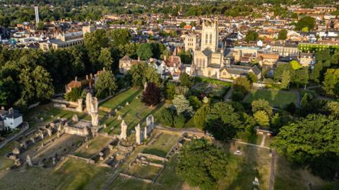 An aerial image of the sand-coloured St Edmundsbury Cathedral, which has a square tower with tall spikes at each corner. In the foreground is are grey stumps which are the ruined masonry of a medieval abbey. The site is surrounded by trees and in the background the buildings of a town centre. 