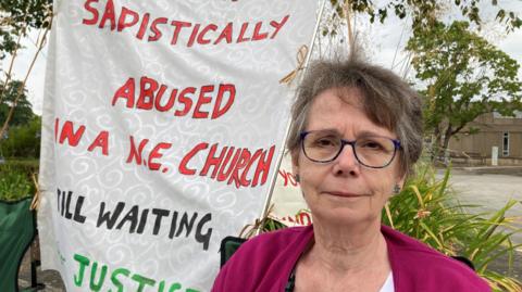 Jenny Read is sitting on a camping chair protesting outside the Church of England's parliament, the General Synod. Behind her is a banner which reads 'sadistically abused in a NE church, still waiting for justice' - although the word "for" is hidden in the fold of the banner. It's written in red, green and black on what looks like embossed, cream furnishing or curtain fabric.