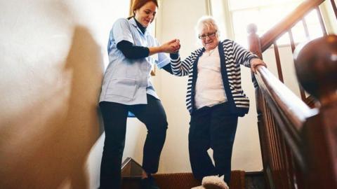 A care worker helps an elderly lady down the stairs.