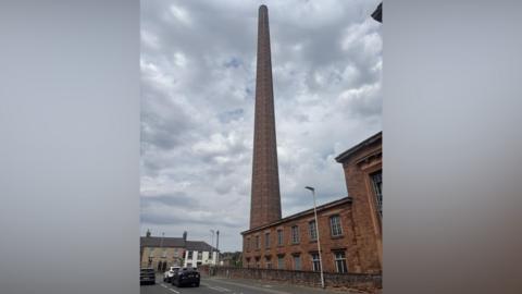 Dixon's Chimney. It is a red brick structure that towers above all other buildings in the area. Several cars are stopped at nearby traffic lights. The sky is filled with clouds.