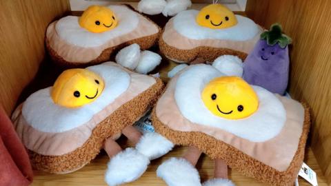 Five Jellycat toys on a shelf. Four of them are egg on toast toys with black eyes and a smiley face on the yolk wearing fluffy white slippers. There is also a purple aubergine with a smiley face.