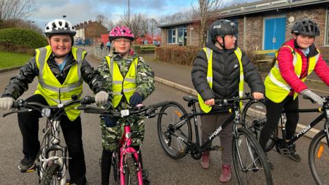 Four children are pictured on their bikes. They re all wearing high-vis jackets over their coats. A boy on the left is wearing a white helmet and smiling. A girl to his right is on a pink bike and wearing a pink and white spotted helmet. Two other boys are wearing black helmets.