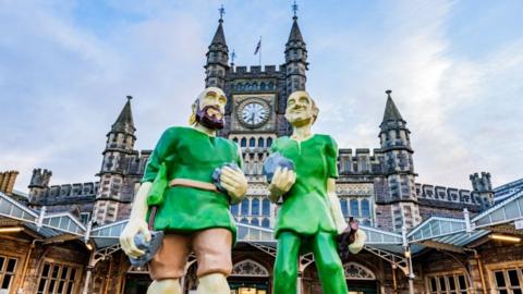 Statue of two mythical giants dressed in green standing outside the entrance to Bristol Temple Meads Station.