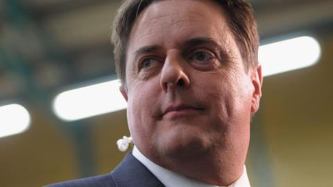 Nick Griffin is wearing a suit jacket and a white collared shirt, looking past the camera. Behind him are bright lights as he stands indoors.