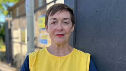 A woman with short dark hair looks at the camera. She is wearing a bright yellow tabard 