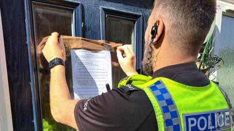 View of the back of a police officer affixing a printed notice to a dark blue front door with brown parcel tape. He has close cropped grey hair and a beard.