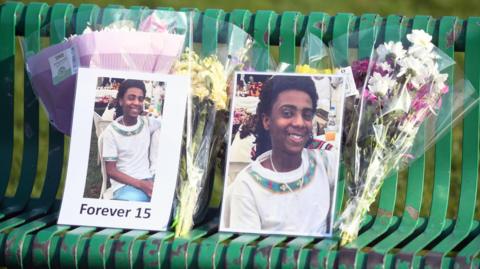 Tributes to Amen on a green bench. There are pictures of him smiling and bunches of flowers.