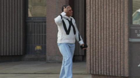 Elizabeth Adeagbo, who is black, has short hair and is wearing a white woollen jumper with a black pattern and jeans, stands outside a brown pebble-dashed court building holding a black mobile phone in her left hand and covering her face with her left. 