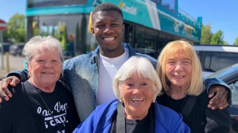 Lynn Crawford, 71, Susie Matthews, 73, Rose Sulley, 74 and Man Like DeReiss photographed next to a Cardiff bus. DeReiss is stood in the middle of the women with his arms around them.