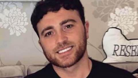 Man who was stabbed to death - a family photograph of Reece Newcombe on a sofa, wearing a black top and smiling with his head tilted to one side.