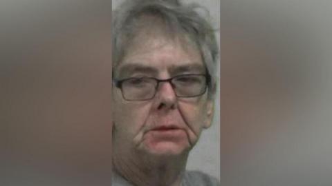 Mugshot of Maria Chambers. She is an older looking lady with messy grey hair and black glasses. Her eyes are half closed.