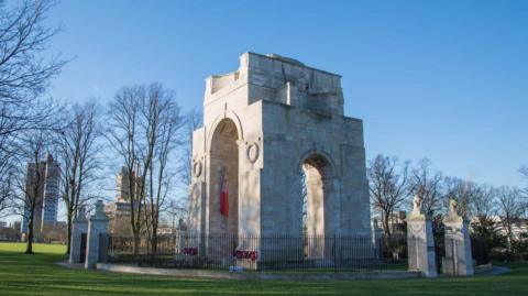 A large memorial archway made of very light coloured stone, surrounded by an ornate iron fence and sited within an area of park land.